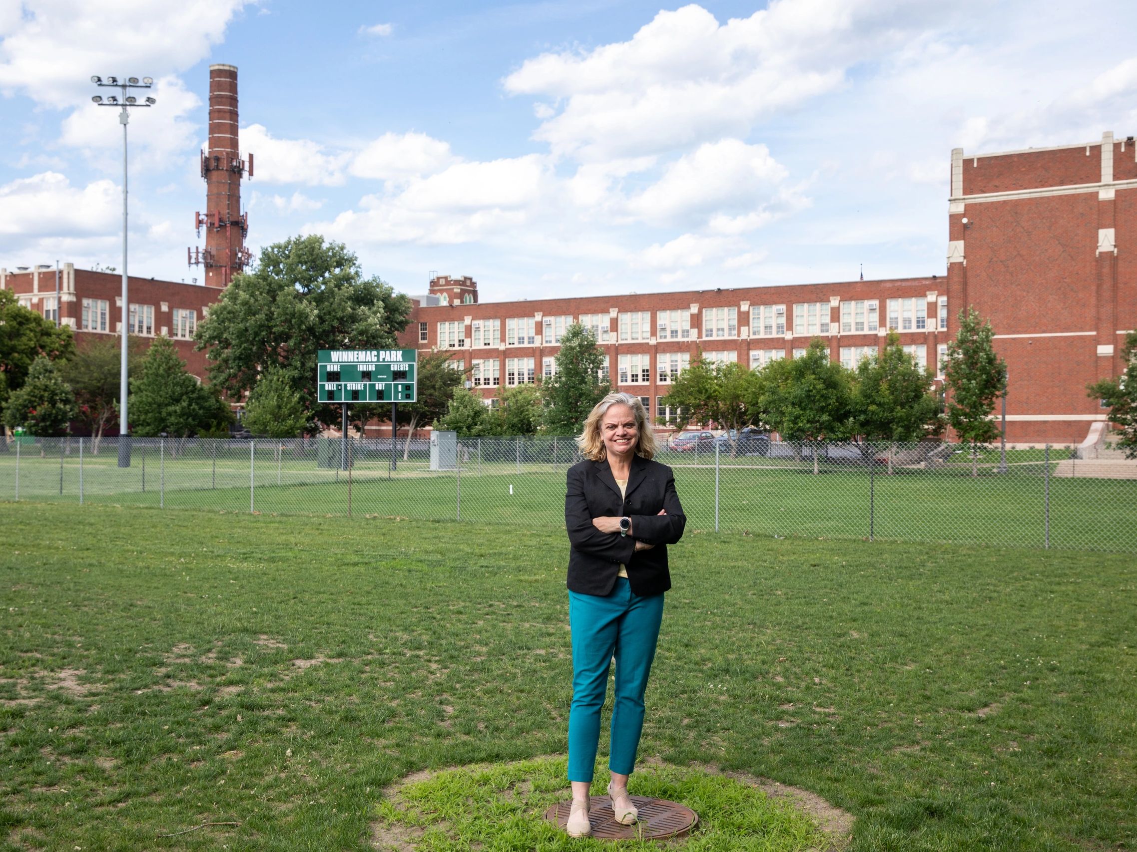 Woman standing on grass field with a school building and scoreboard in the background.