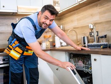 A technician is fixing a dishwasher in a stylish kitchen while smiling at the camera.