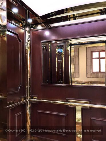 Vista a 45 grados y pared de fondo con espejo de un elevador de madera natural y acero dorado