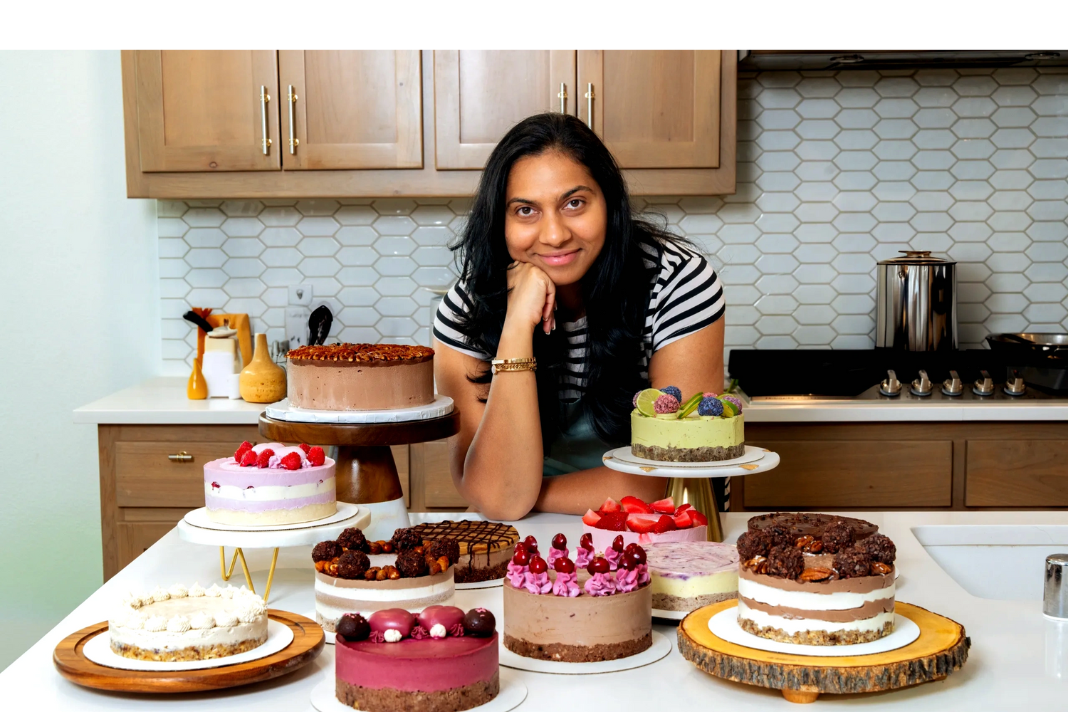 Woman smiles behind a variety of colorful cakes in a modern kitchen.
