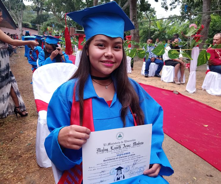A student in a blue graduation gown smiles and shows her new high school diploma.