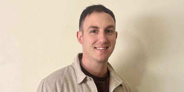 Smiling man in beige shirt against plain wall.