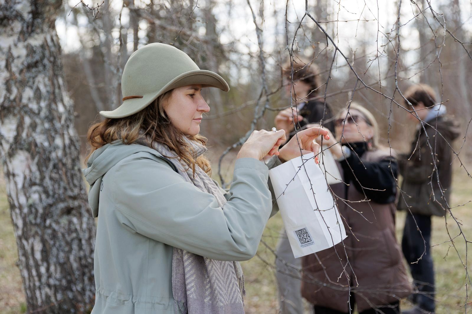 Woman hanging a small white paper bag on tree branches outdoors.