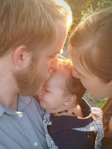 Parents tenderly kiss their baby in a sunlit garden.