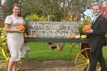 Two people holding pumpkins stand beside a 'Welcome to Dickinson' sign on a decorated wagon.
