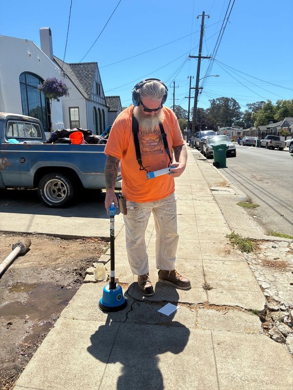 Man using ground scanning equipment on a sidewalk under clear blue sky.