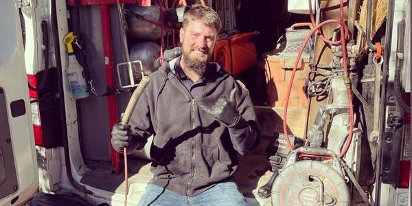 A worker sitting in a van surrounded by plumbing tools, giving a thumbs up.