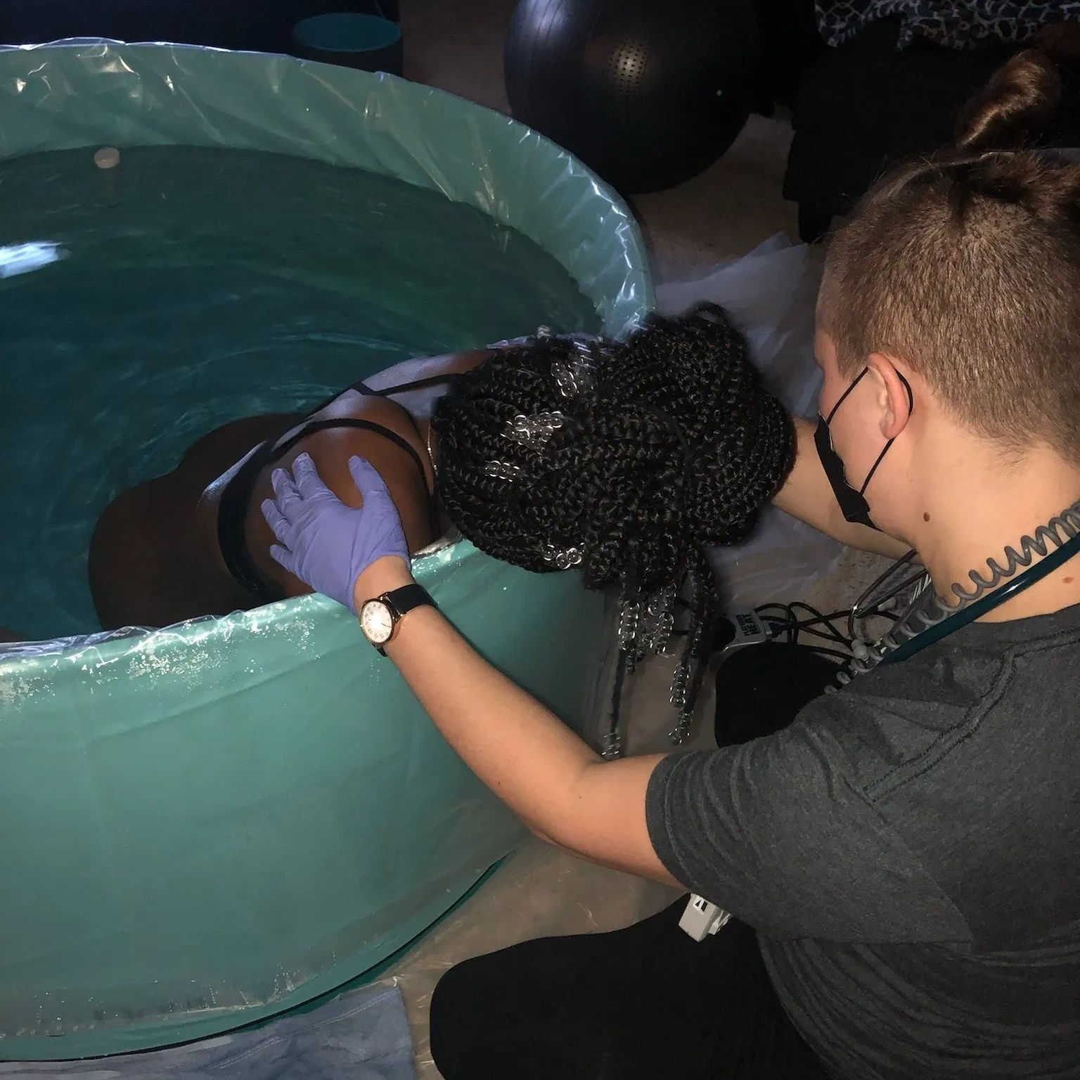 A woman in a birthing pool supported by a caregiver wearing gloves and a mask.