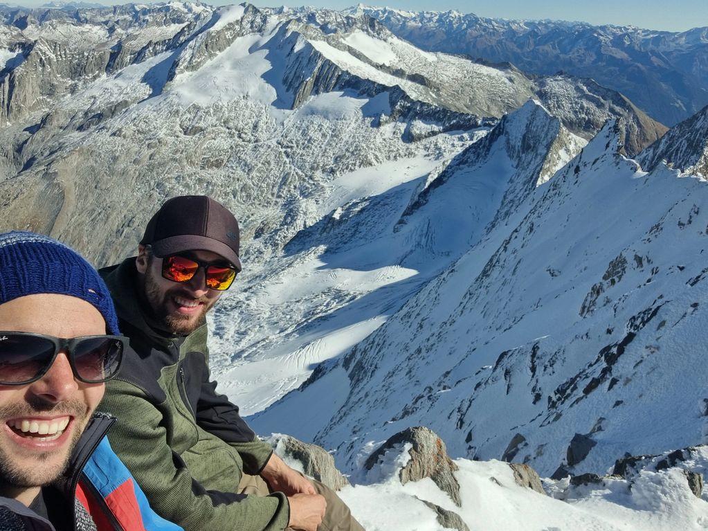Two men smiling on a snowy mountain peak with expansive mountain views.