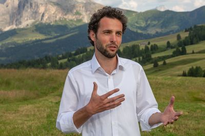 Man in white shirt gesturing while speaking outdoors in a mountainous landscape.
