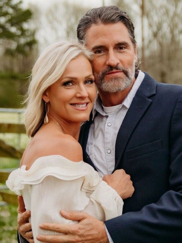 A couple embraces outdoors with warm smiles, dressed elegantly.