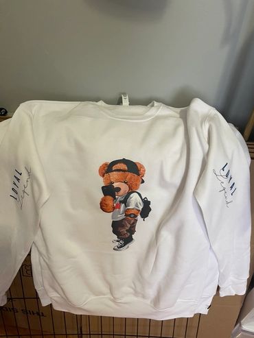White sweatshirt with a teddy bear graphic holding a phone and wearing streetwear.