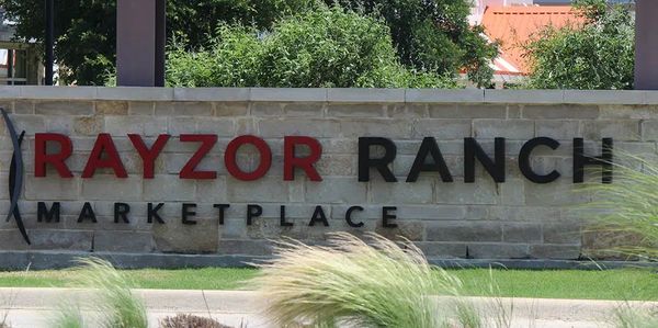 Stone sign for Rayzor Ranch Marketplace with greenery in the background.