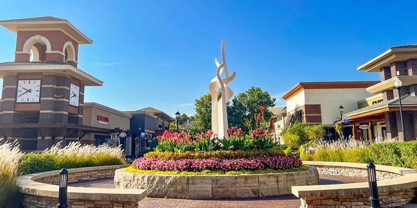 A vibrant shopping plaza with a central flowerbed and modern sculpture under a clear blue sky.