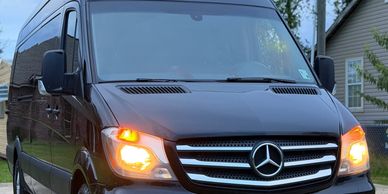 Black Mercedes van with headlights on in a suburban driveway.
