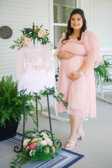 Mother-to-be standing on porch next to baby shower welcome sign.