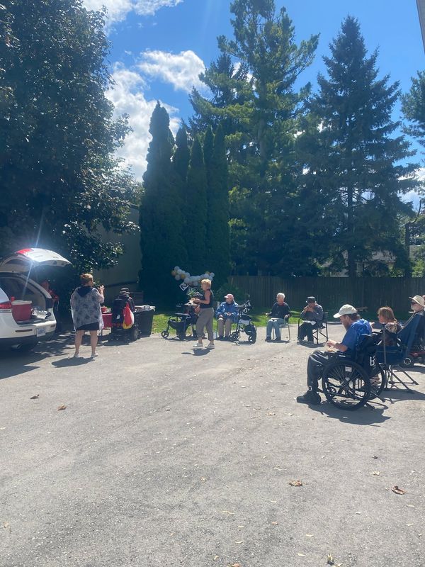 Outdoor gathering of elderly people, some in wheelchairs, enjoying a sunny day with caregivers.