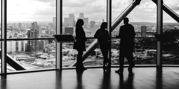 Silhouettes of three people standing by large windows overlooking a cityscape.