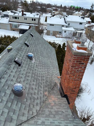 Snow-covered rooftops with a brick chimney and skylights in a suburban neighborhood.