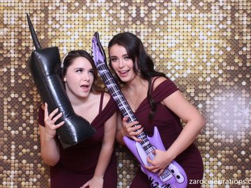 Two guests are having fun in the photo booth. Gold sequin backdrop and inflatable props.