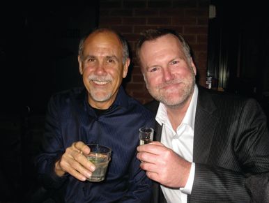 Two men smiling and toasting with drinks indoors at night.