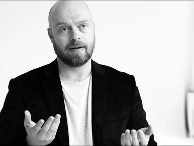 A man in a black blazer gestures while speaking thoughtfully.