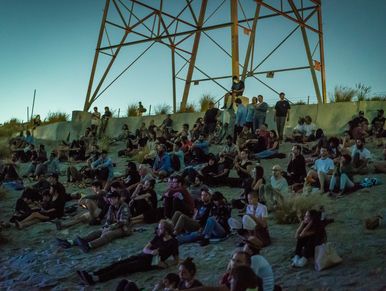 Large group of people sitting outdoors under a metal structure at dusk.
