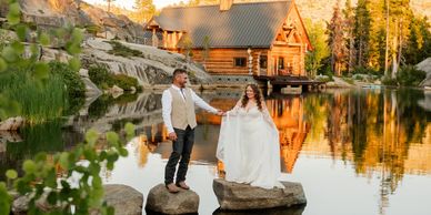 Couple in wedding attire holding hands on rocks by a serene lake with a cabin backdrop.