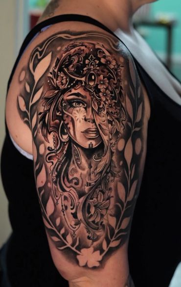 Intricate black and gray tattoo of a woman's face adorned with floral and ornamental designs on upper arm in a realistic style. Realistic woman tattoo with flowers done at Artistic Minds Tattoo. We're the best Conroe tattoo shop, the best tattoo Houston shop, and best tattoo shop near the woodlands.