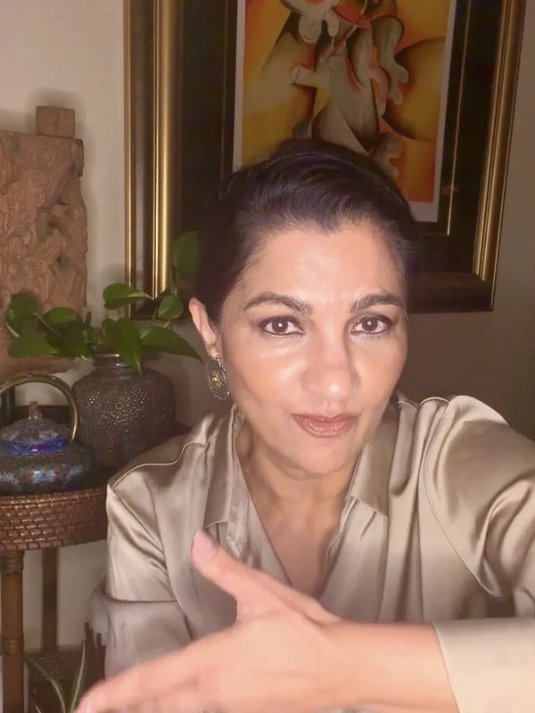 A woman in a satin blouse poses indoors with framed art and plants in the background.