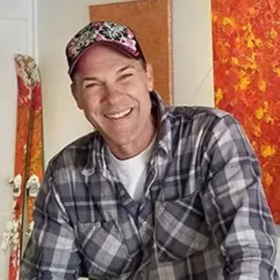 Smiling man wearing a plaid shirt and decorative cap indoors.