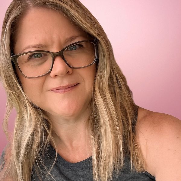 Woman with glasses and blonde hair smiling against a pink background.