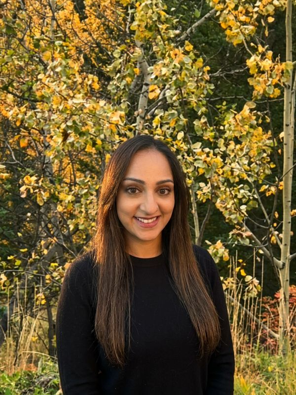 A smiling woman with long hair stands outdoors in front of autumn trees.