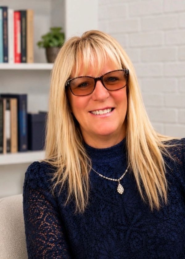 Smiling woman with blonde hair and glasses wearing a navy lace top and a pearl necklace.