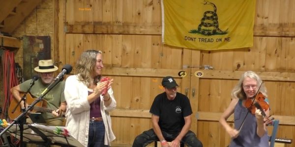 A group of musicians playing and clapping in a rustic wooden room with a "Don't Tread On Me" flag.