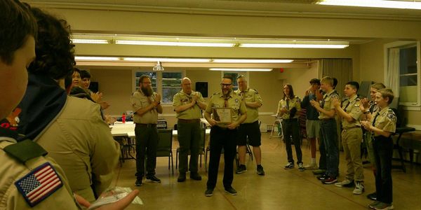 Boy scouts and leaders applauding a man holding an award in a room.