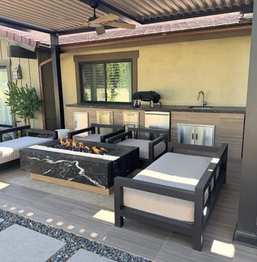 Modern outdoor patio with fire pit, lounge chairs, and kitchenette under a covered roof.