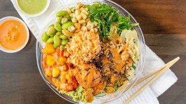 Colorful poke bowl with edamame, seaweed, mango, and spicy sauce with dipping sauces and chopsticks.