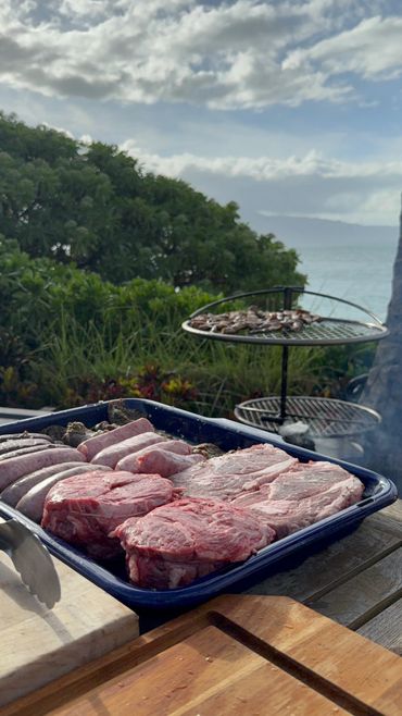 Raw meats and sausages prepared for grilling outdoors with a scenic nature background.