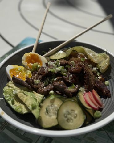 Bowl of beef, soft-boiled eggs, avocado, cucumber, and radishes with chopsticks.
