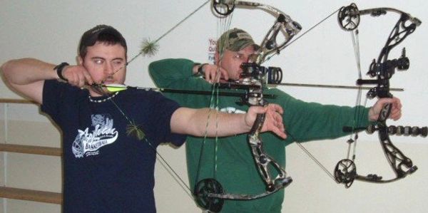Two men aiming with compound bows indoors, preparing to shoot arrows.