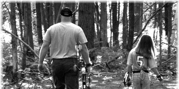 A man and a child walk through a forest carrying bows for archery.