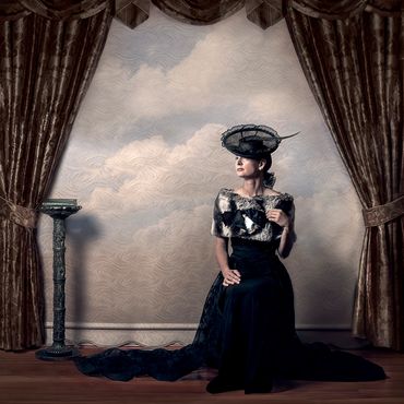 Fine art;female portrait;black hat feather;pedestal;book;long golden drapes;clouds on cream blinds. 