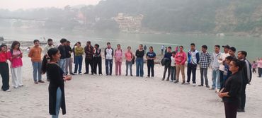 Group of people standing in a large circle by the riverside with hills in the background.