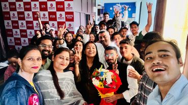 A large group of happy people posing for a selfie with a bouquet of flowers indoors.