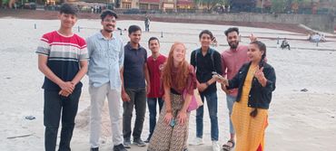 A group of friends posing happily on a sandy beach with smiles and peace signs.