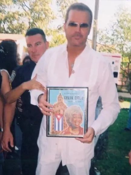 Man in white holding a framed Celia Cruz poster outdoors.