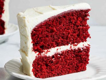 Slice of red velvet cake with creamy white frosting on a white plate.