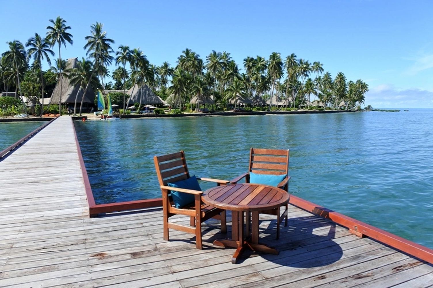 Wooden chairs and table on a pier overlooking a tropical island with palm trees.