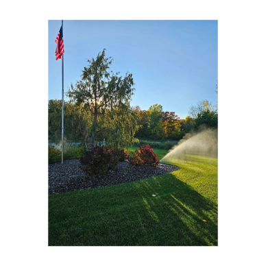 A sprinkler waters a green lawn near an American flag on a sunny day.